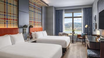 A hotel room with two beds, a desk, a chair, a view of the outdoors, and a patterned wall.