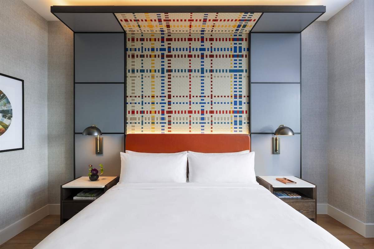 Habitación de hotel con una cama grande, dos mesitas de noche, una lámpara de pared y un diseño geométrico colorido en la pared.