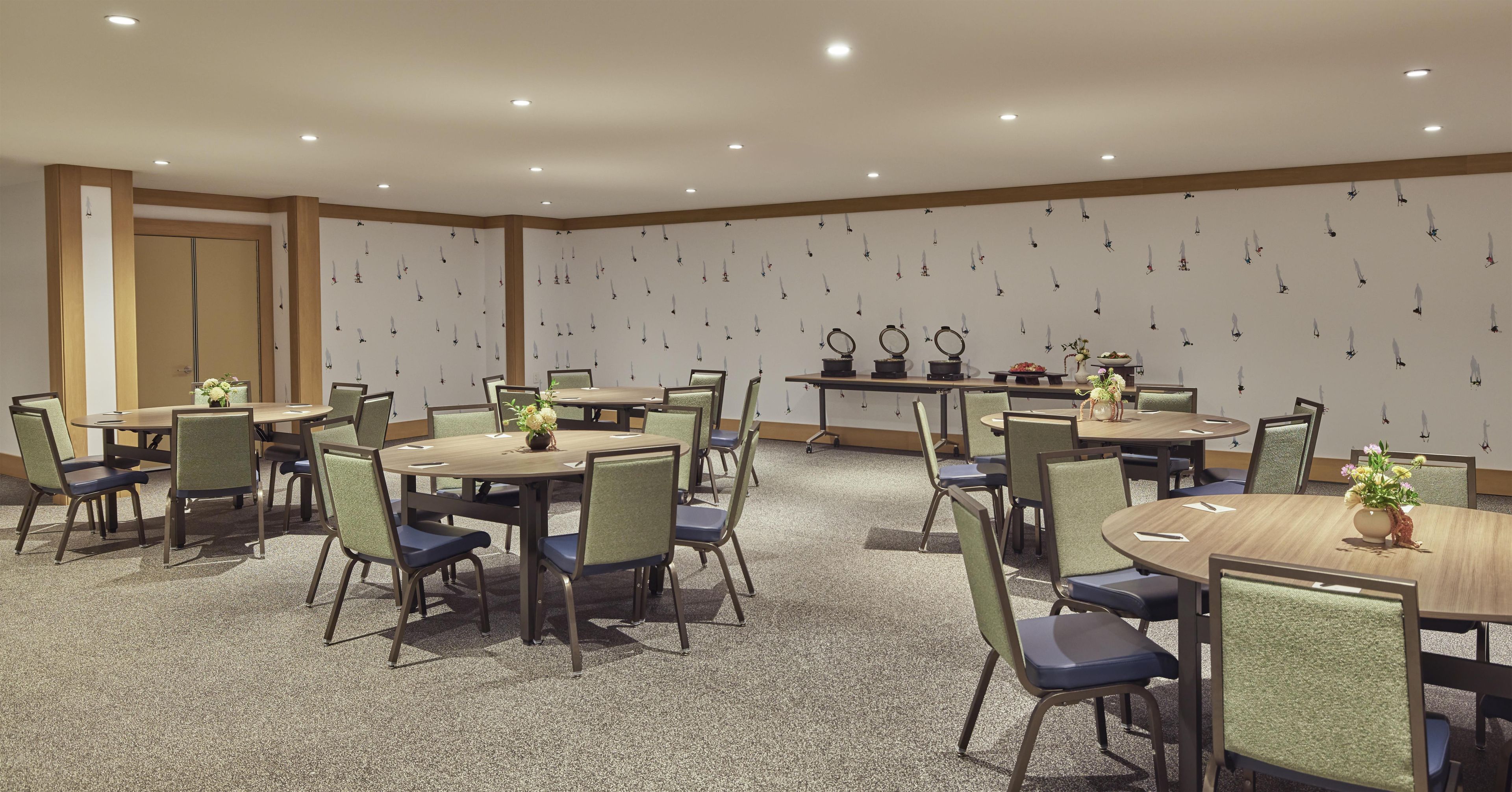 Un salón de conferencias con varias mesas y sillas, equipado con iluminación en el techo y un muro decorativo.