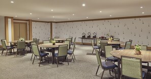 Un salón de conferencias con varias mesas y sillas, equipado con iluminación en el techo y un muro decorativo.
