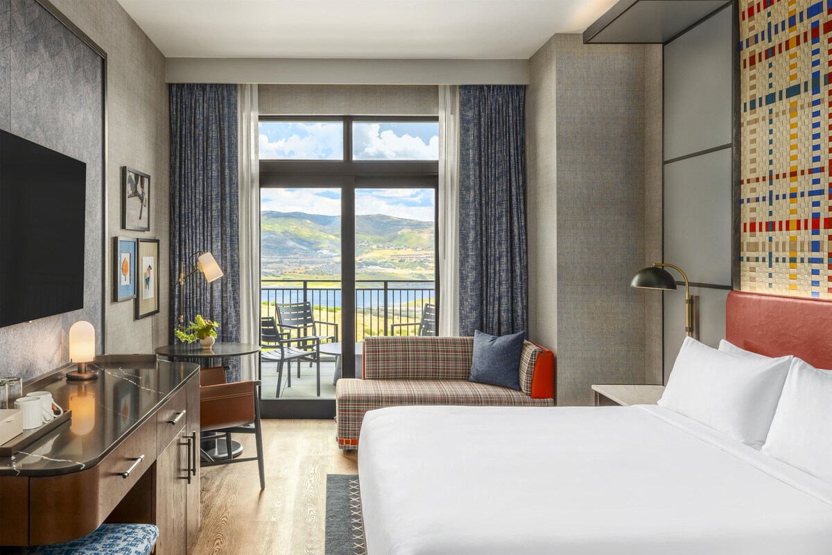 A hotel room with a bed, a desk, a chair, a sofa, and a balcony with a view of the countryside.