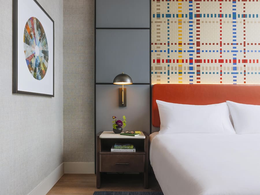 Una habitación de hotel con una cama, una mesita de noche, una obra de arte enmarcada, una lámpara y un papel tapiz estampado.