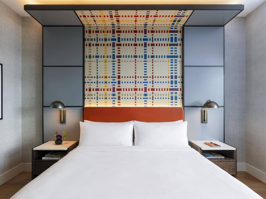 Habitación de hotel con una cama grande, dos mesitas de noche, una lámpara de pared y un diseño geométrico colorido en la pared.
