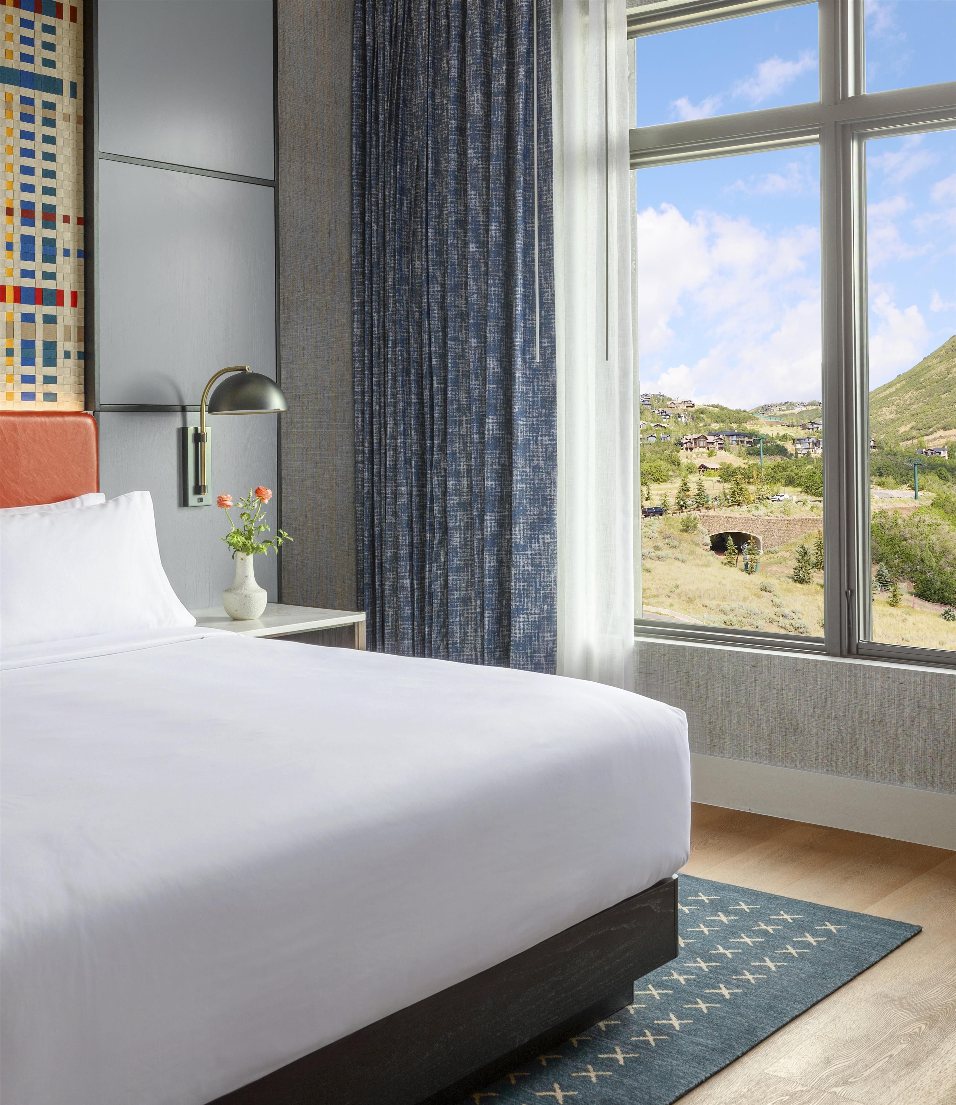 Een hotelkamer met een groot bed, een nachtkastje met een lamp, een vaas met bloemen, en uitzicht op een bergachtig landschap door een raam, plus een kamerbreed tapijt met dessin.