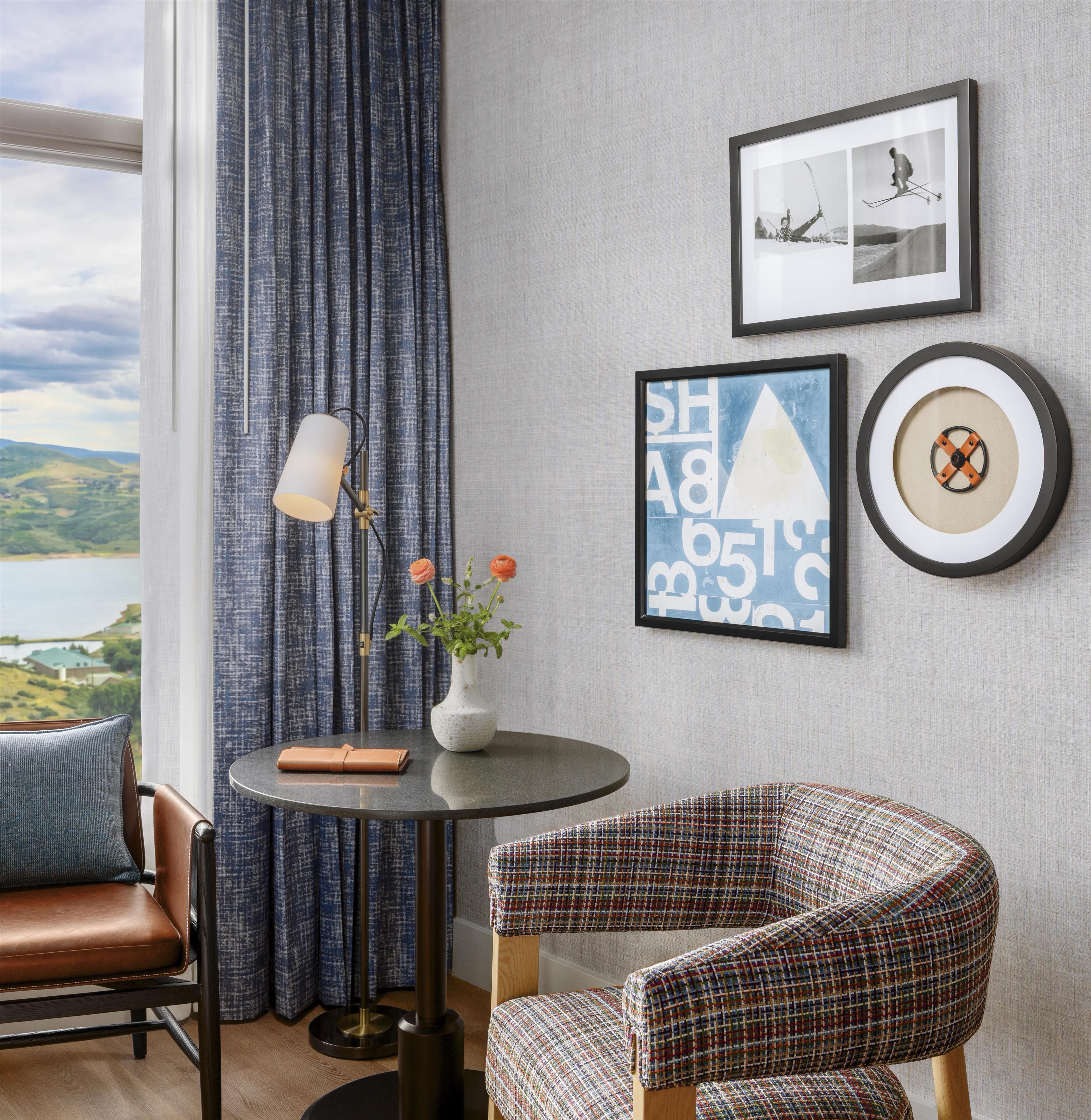A room with a view of a lake, a round table with a vase of flowers, a patterned armchair, and framed artwork on the wall.
