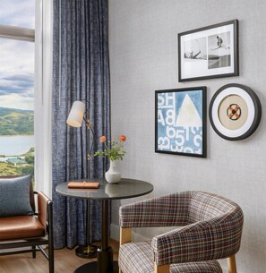 Una habitación con vistas a un lago, una mesa redonda con un florero, un sillón estampado y obras de arte enmarcadas en la pared.