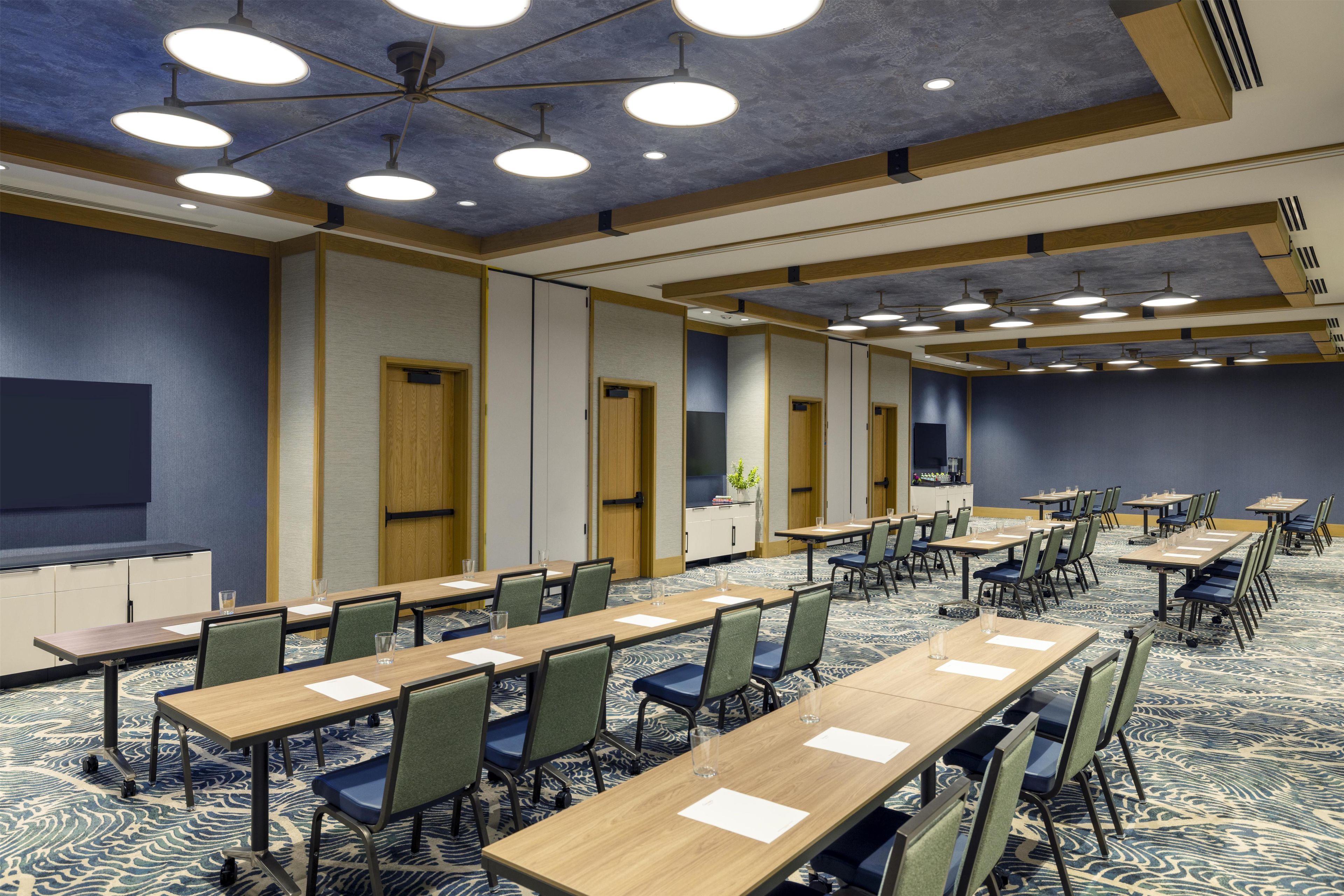 Un salón de conferencias con varias mesas y sillas, una pantalla grande e iluminación empotrada en el techo.
