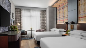 Habitación de hotel con cama, mesitas de noche, escritorio, silla, mesita pequeña y ventana con cortinas.