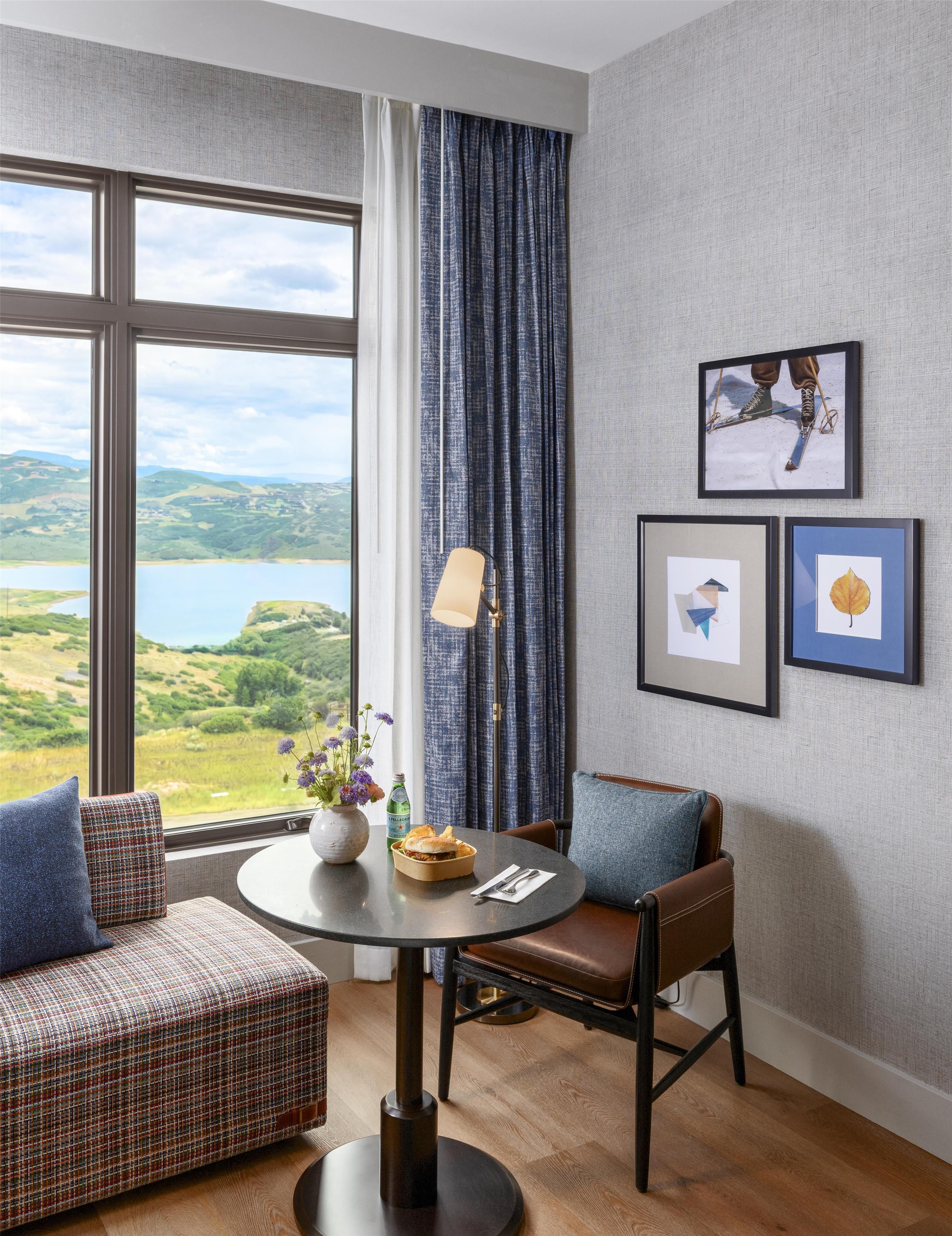 A room with a view of a lake, a dining table with a vase of flowers, and two framed pictures on the wall.