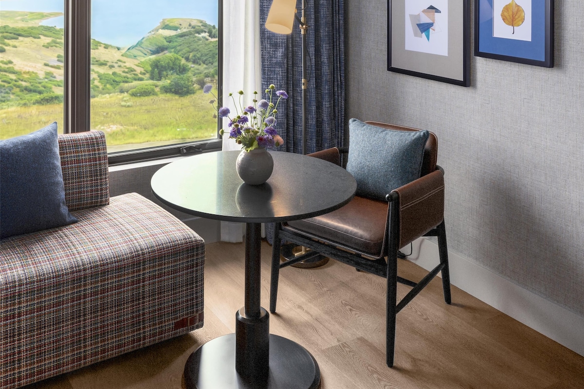 Una habitación con un ventanal grande, una mesa redonda, una silla, un sofá y cuadros en la pared.