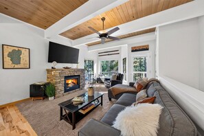 Ein modernes Wohnzimmer mit Kamin aus Stein, Deckenventilator und großem Fenster mit Blick auf eine Terrasse.