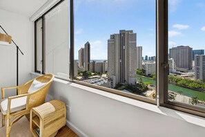 A room with a large window, a wicker chair, and a view of a cityscape with tall buildings and a river. - Oceanview Condo + Pool + Gym | 10-Min Walk to Beach | Coastal Getaway Waikiki (Honolulu)