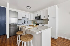 A modern kitchen with white cabinetry, a stainless steel refrigerator, a microwave, and a marble island with stools. - Oceanview Condo + Pool + Gym | 10-Min Walk to Beach | Coastal Getaway Waikiki (Honolulu)