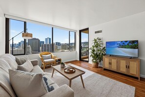 A modern living room with a large window, a sofa, a coffee table, a TV, and a plant. - Oceanview Condo + Pool + Gym | 10-Min Walk to Beach | Coastal Getaway Waikiki (Honolulu)