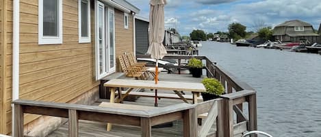 A wooden deck with outdoor seating and a covered umbrella, overlooking a canal with boats.