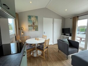 A modern living room with a round dining table, wooden chairs, a grey sofa, a flat-screen TV, and a painting on the wall.