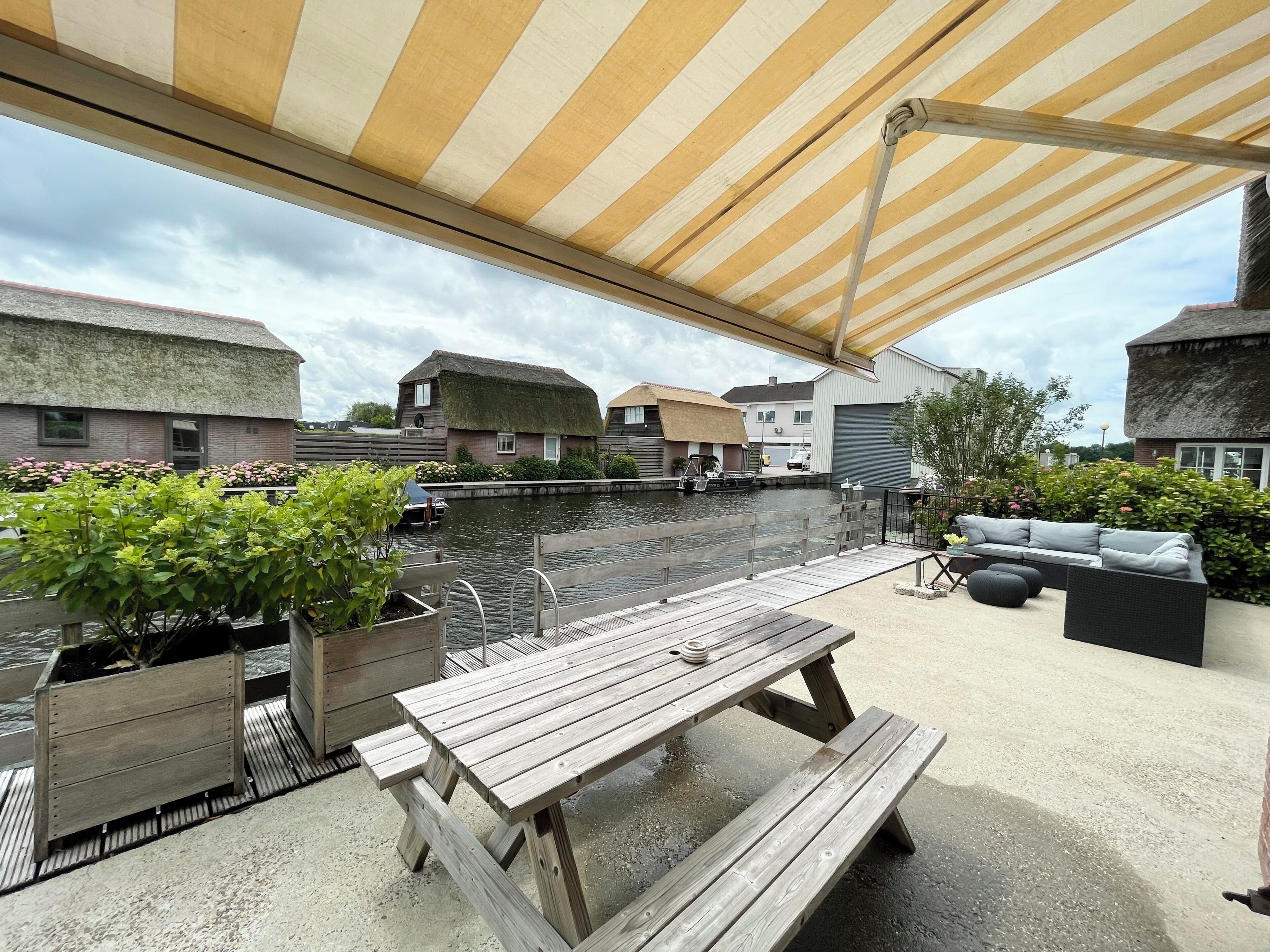 A covered outdoor area with a wooden table and benches, a small boat docked by the water, and a thatched-roof building in the background.