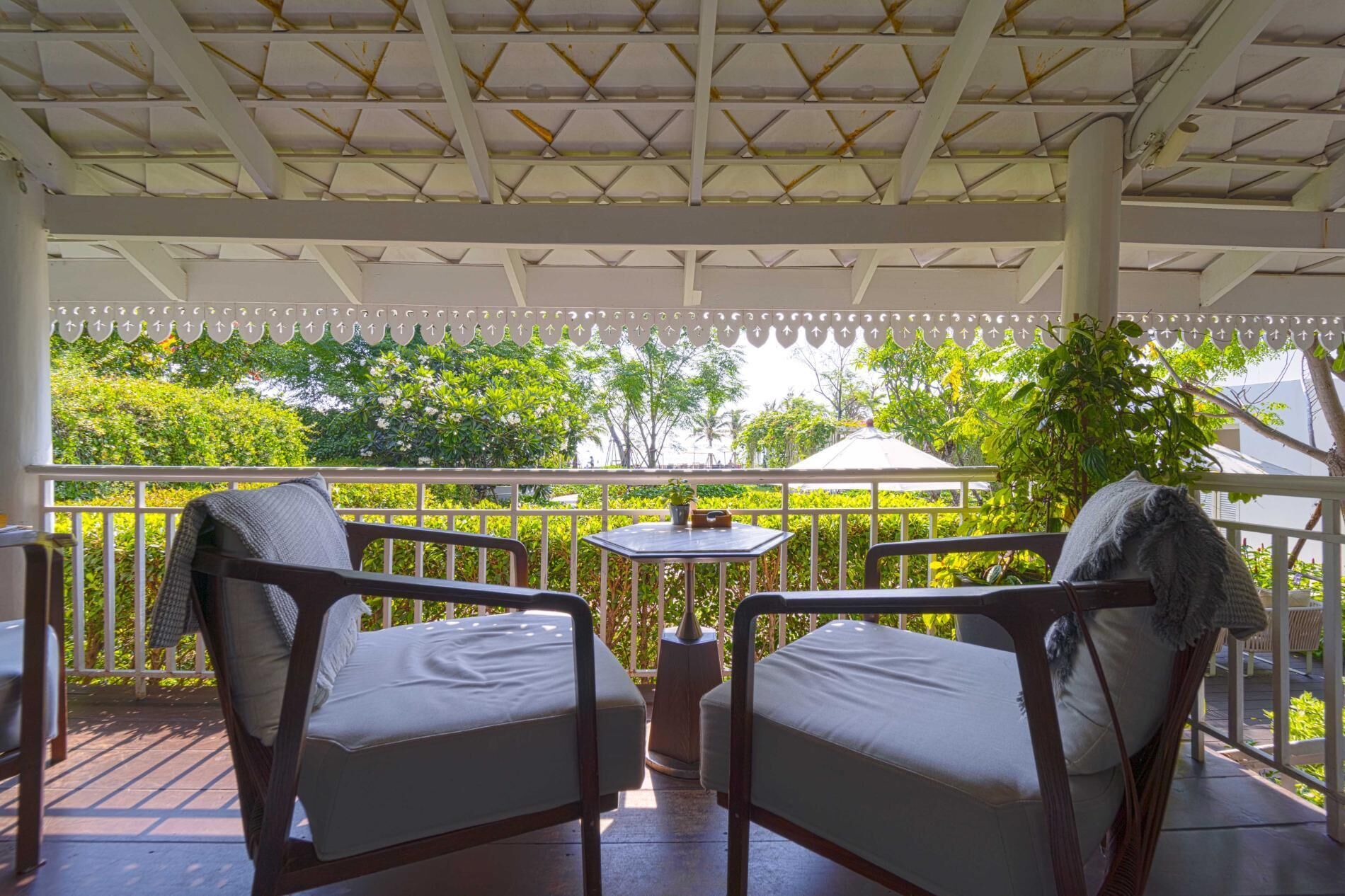 A covered outdoor seating area with two chairs and a table, surrounded by greenery and a white railing.