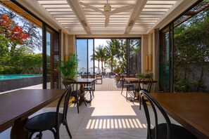 Ein Außenbereich zum Essen mit Tischen und Stühlen, einem Deckenventilator und Blick auf das Meer.