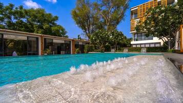 A swimming pool with a waterfall feature, surrounded by trees and modern buildings.