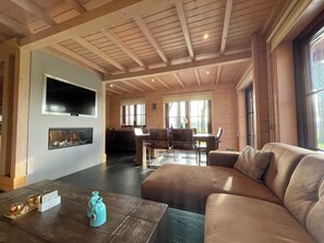 A spacious living area with a fireplace, a large sofa, a dining table, and a kitchen in the background. - 4-person holiday home on a small-scale holiday park with infrared sauna and beautiful views in a quiet area (Den Ham)