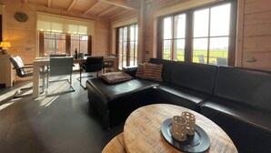 Une pièce spacieuse avec un plafond en bois, un canapé en cuir noir, une table à manger avec des chaises et de grandes fenêtres offrant une vue sur l’extérieur.