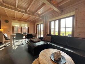 Ein geräumiger Raum mit Holzboden, einem schwarzen Ledersofa, einem Esstisch mit Stühlen und großen Fenstern mit Blick ins Freie.