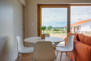 Ein Zimmer mit rundem Tisch, zwei weißen Stühlen, einem orangefarbenen Sofa und Blick auf ein Gebäude durch ein großes Fenster.