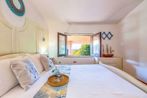 Ein Schlafzimmer mit Bett, Kissen, einem Nachttisch mit einem Schiffmodell, einem Fenster mit Blick auf Grünflächen und einer Wanddekoration.