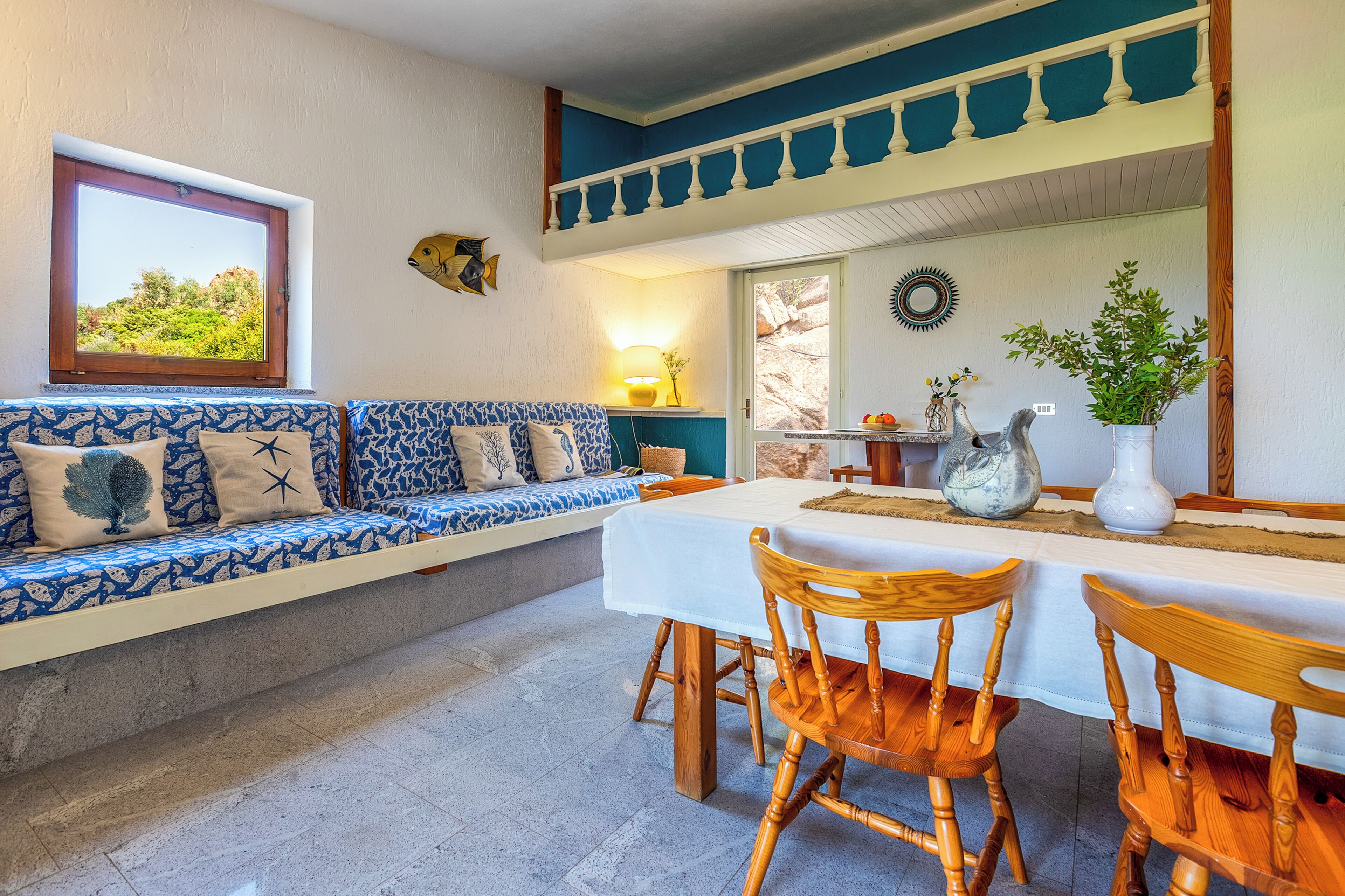 A room with a sofa, wooden chairs, a table, a window with a view, and a balcony.