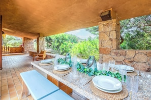 An outdoor dining area with stone walls, a stone fireplace, and a view of greenery. - COSY VILLA Sea View and Garden (Costa Paradiso)
