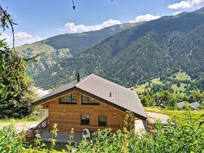 Uma cabana de madeira com telhado inclinado, duas janelas e chaminé, cercada por vegetação exuberante e montanhas.