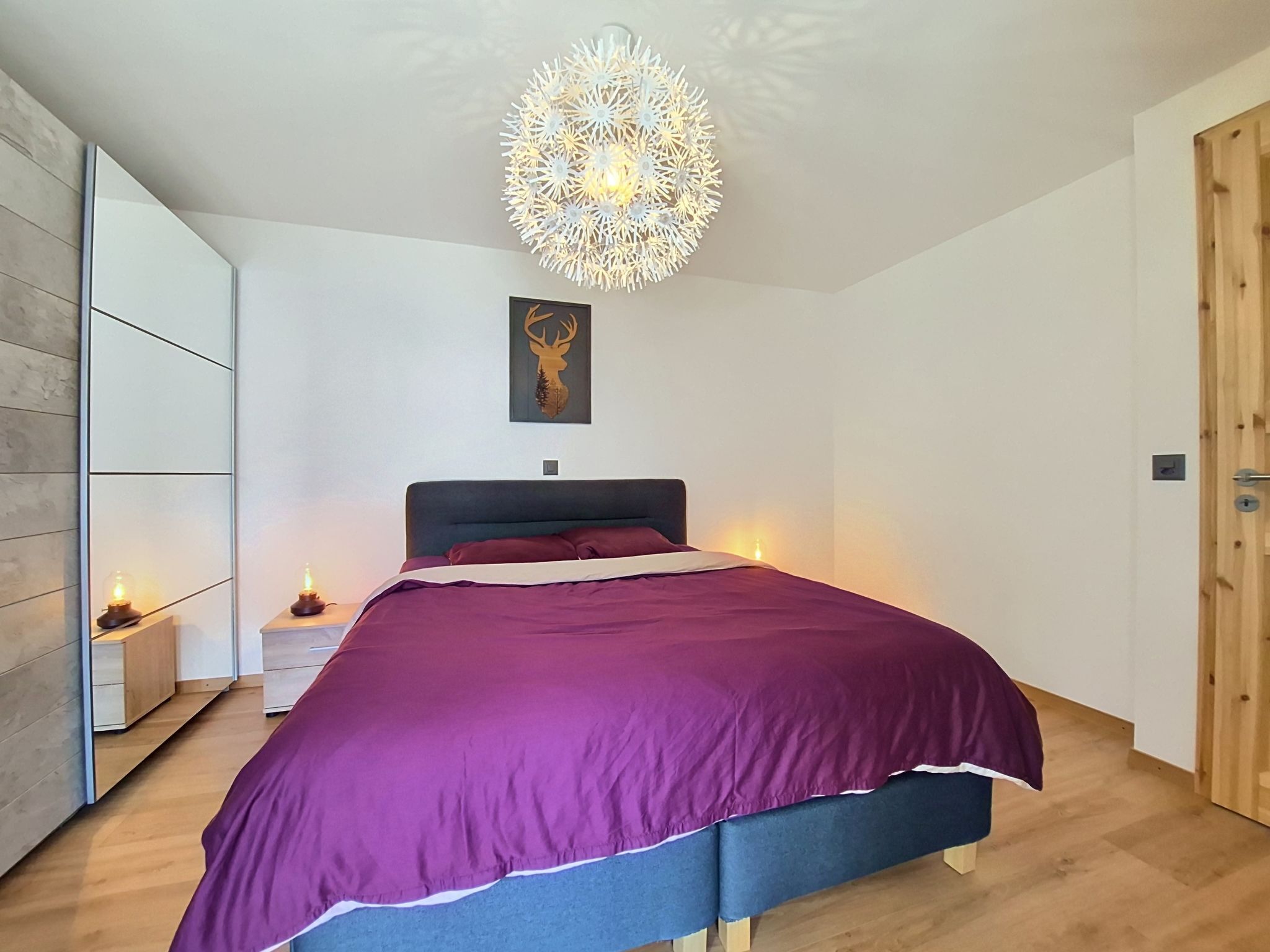 A bedroom with a bed, a wall-mounted light fixture, a framed deer head artwork, and a wooden door.