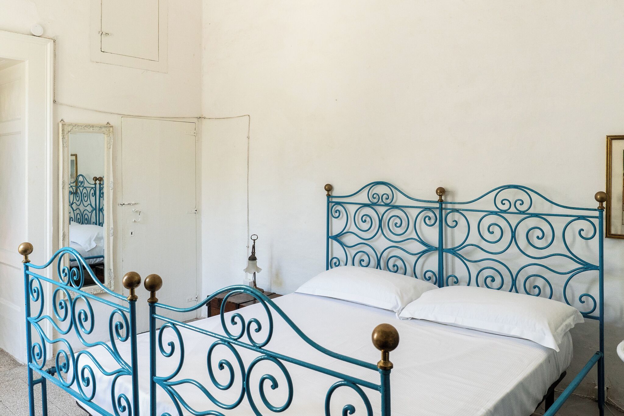 A bed with a blue metal frame and white bedding, a white door, and a mirror with a decorative frame.