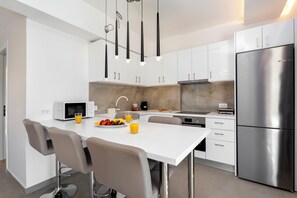 A modern kitchen with white cabinets, a stainless steel refrigerator, a white island with bar stools, and pendant lighting. - Orama Sunset | Emily Villa (Rhodes)