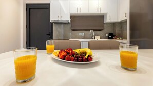A modern kitchen with white cabinets, a dark door, and a dining table with fruit and juice glasses. - Orama Sunset | Joana Villa (Rhodes)