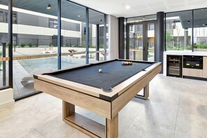 Et billardbord med træramme og sort filtdug, placeret i et rum med store vinduer, der giver udsigt til et udendørs poolområde og en bygning i baggrunden.