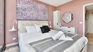 A bedroom with a large bed, a nightstand, a dresser, a mirror, and a wall art piece. - Newcastle Views at One Apartments (Newcastle West)