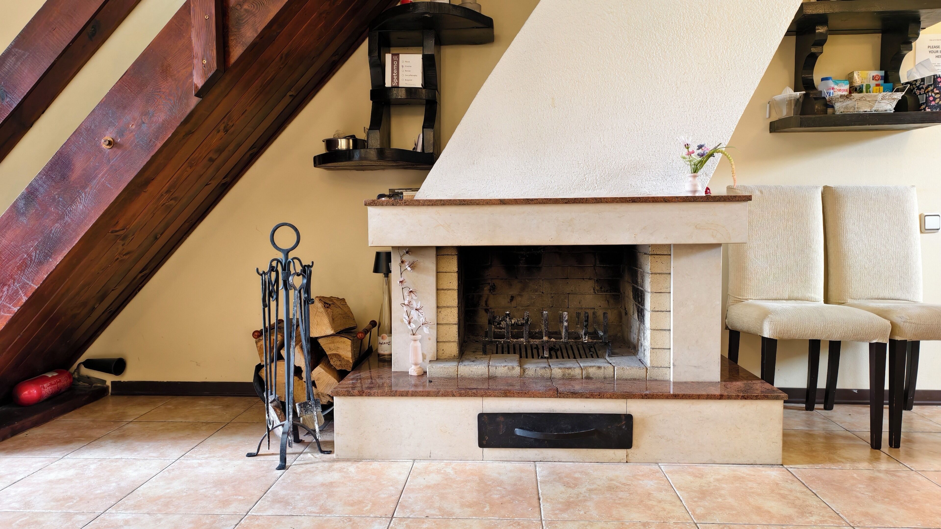 A fireplace with a stone surround, a chair, a shelf with items, and a wooden beam structure.