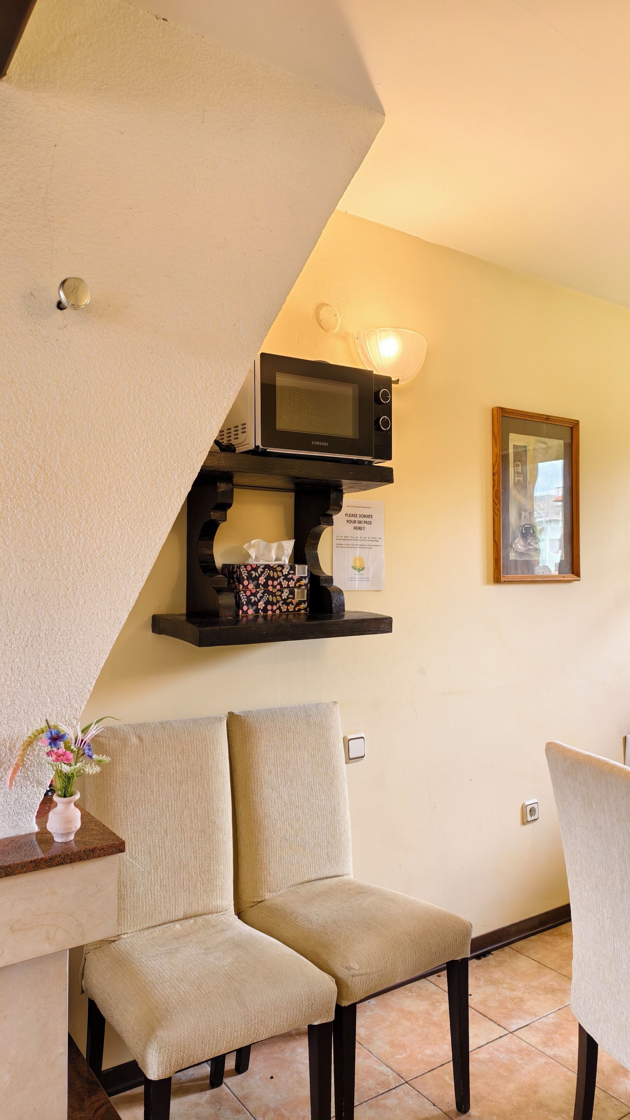 Une pièce avec une télévision posée sur une étagère, une chaise, une petite table sur laquelle se trouve un vase de fleurs, et un tableau encadré accroché au mur.