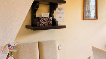 Une pièce avec une télévision posée sur une étagère, une chaise, une petite table sur laquelle se trouve un vase de fleurs, et un tableau encadré accroché au mur.