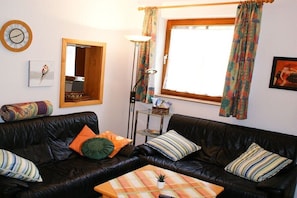 Ein Wohnzimmer mit schwarzem Ledersofa, einem kleinen Tisch, einer Stehlampe, einem Fenster mit Vorhängen, einer Uhr und einem gerahmten Bild an der Wand.