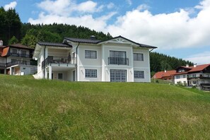 Ein großes, mehrgeschossiges Gebäude mit Balkonen, gelegen auf einem grasbewachsen Hügel mit Wald im Hintergrund.