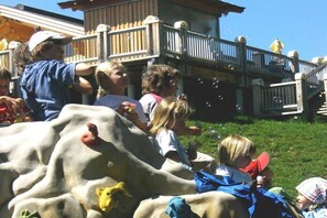 Eine Gruppe Kinder spielt mit Seifenblasen in einem Freigelände mit Holzzaun und einem Gebäude im Hintergrund.