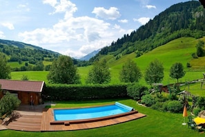 Ein Schwimmbad, umgeben von einer Holzterrasse, einer Holzhütte und einer üppigen, grünen Landschaft mit Hügeln und Bäumen.