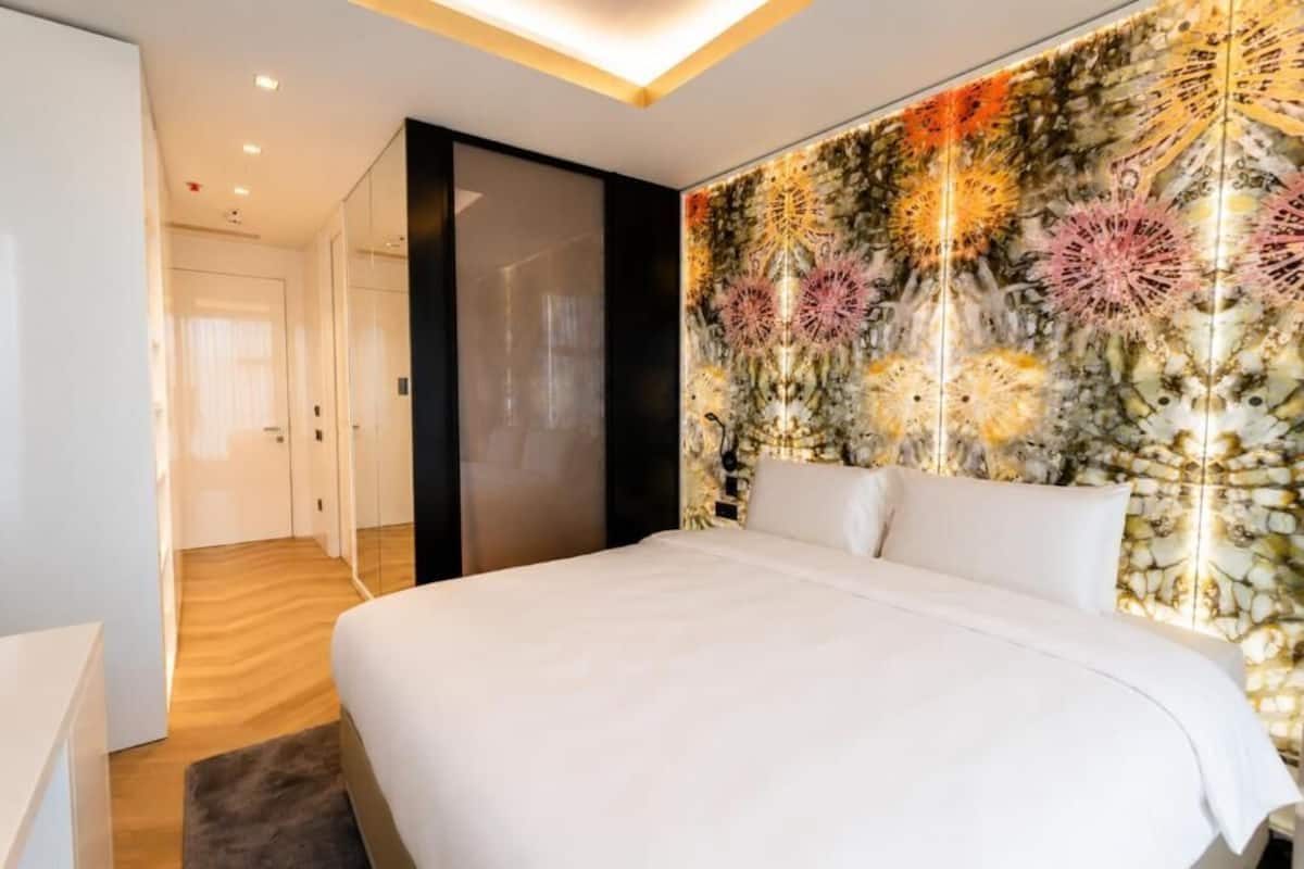 A modern hotel room with a large bed, a decorative wall, and a hallway leading to a door.
