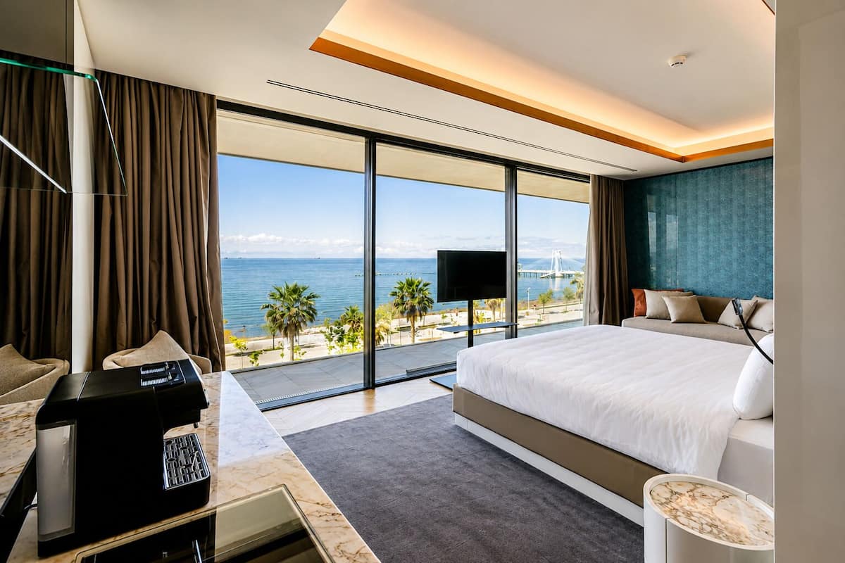 A modern hotel room with a large bed, a desk with a coffee machine, a TV, and a view of the sea.