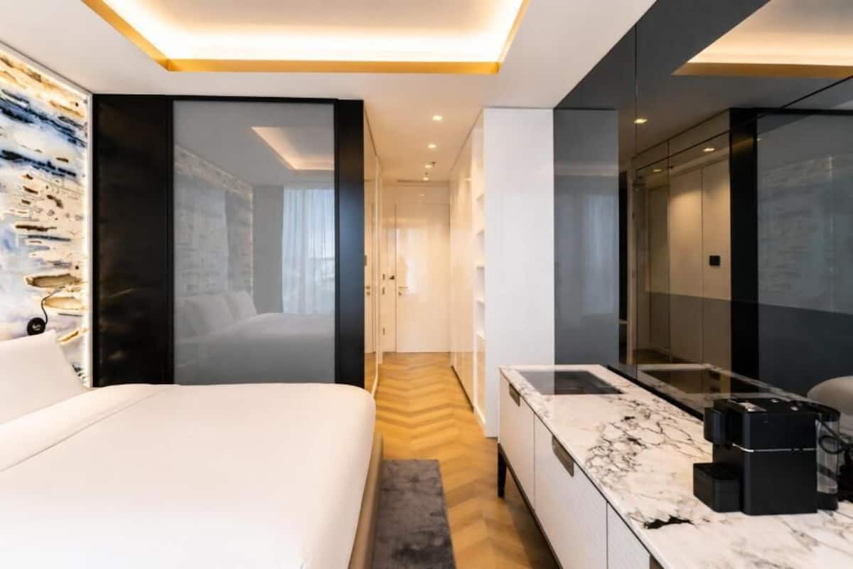 A modern hotel room with a bed, a marble bathroom vanity, and a hallway leading to a bathroom.