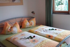 Ein Bett mit blumigem Bettzeug und Kissen, ein Metall-Kopfteil, ein Fenster mit Blick auf Bäume und ein an der Wand befestigter Lichtschalter.