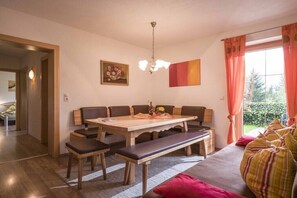Ein Essbereich mit einem Holztisch, Bänken und einer Sitzbank. An der Wand hängt ein Gemälde, eine Chandelieber ist angebracht, und ein Fenster ist mit Vorhängen drapiert.
